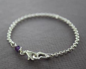 May include: A delicate silver bracelet with a small, round chain design. The bracelet features a lobster clasp closure and a single, small purple gemstone. The jewellery is displayed on a neutral background.