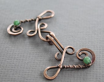 May include: A copper wire jewellery clasp with two green gemstone accents. The clasp is in the shape of a stylised letter 'S' with a twisted wire design.