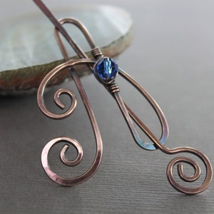 May include: Copper-toned hair stick with a swirling design and a blue crystal accent. The metal is shaped into elegant curves and spirals, with a central blue crystal adding a pop of color. A stylish hair accessory.