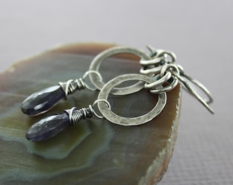 May include: Silver earrings with a hammered finish. Each earring has a dark purple, faceted gemstone hanging from a silver hoop. The gemstones are wrapped with silver wire, and the earrings have a hook closure.