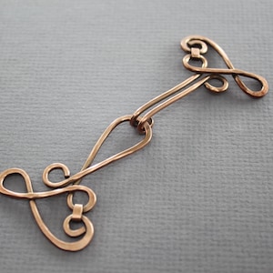 May include: A copper-coloured metal clasp with a decorative design. The clasp is made of two pieces that are connected by a loop in the middle. The design is a series of swirls and loops that create a unique and intricate pattern.