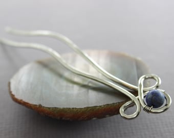May include: Silver hair stick with a blue gemstone at the end. The stick is curved and has a decorative loop at the end.