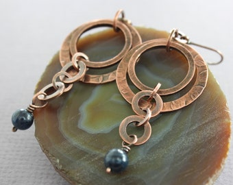 May include: Copper-coloured earrings with a layered design. Each earring features three textured rings connected by small links, with a dark blue bead dangling at the bottom. The earrings are displayed on a green and brown stone.
