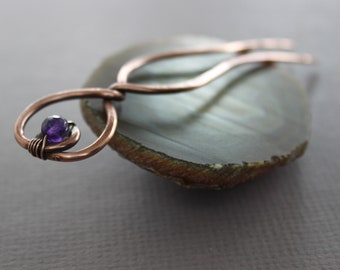 May include: A copper wire hair clip with a purple amethyst gemstone. The clip is shaped like a figure eight and is resting on a piece of agate.