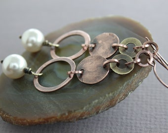 May include: A pair of earrings with a copper and brass design. The earrings feature a large pearl, two copper circles, and several smaller brass circles. The earrings are displayed on a green and brown agate slice.