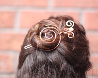 May include: A copper hair stick with a spiral design and a small pearl accent. The stick is holding a bun of brown hair.