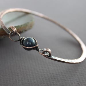 May include: A hammered copper bangle bracelet with a dark blue gemstone wrapped in copper wire.