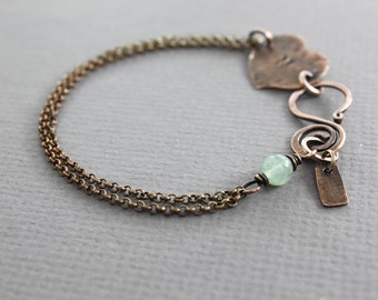 May include: A delicate bracelet featuring a double chain, a heart charm, a green stone bead, and a small tag charm. The bracelet is made of copper and has a rustic, vintage look.