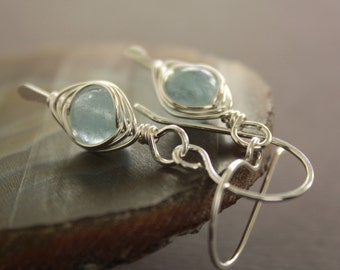 May include: A pair of handcrafted earrings with light blue, oval gemstones set in a silver wire design. The earrings have a delicate, openwork style with a hook closure. The gemstones are approximately 1.3 cm in length.