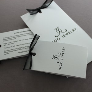 May include: Two white cards with black text and a black ribbon. The top card has the text "Jewelry Care Instructions" and instructions on how to care for jewelry. The bottom card has the text "Ingo Jewelry" in a stylized font.