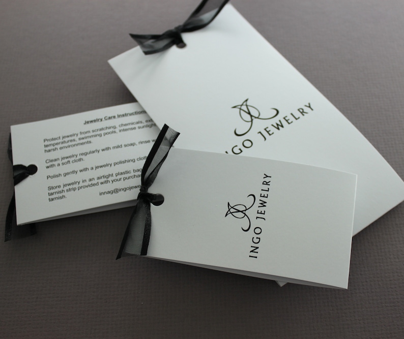 May include: Two white cards with black text and a black ribbon. The top card has the text "Jewelry Care Instructions" and instructions on how to care for jewelry. The bottom card has the text "Ingo Jewelry" in a stylized font.