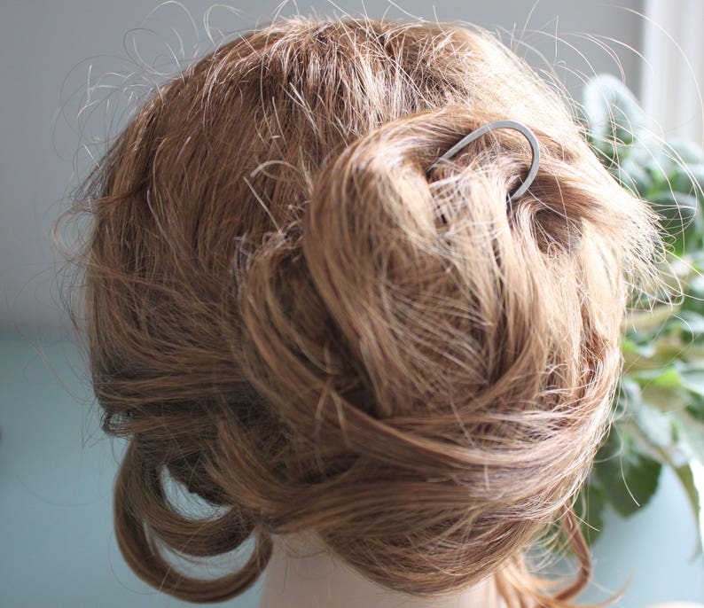 May include: A silver hair pin with a simple curved design, used to hold a bun of light brown hair.