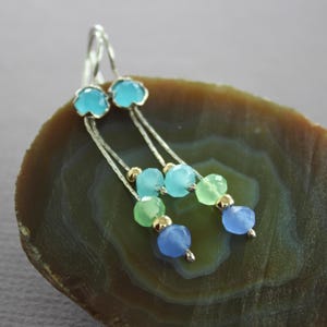 Peut inclure: Boucles d'oreilles pendantes en argent avec une perle supérieure bleu clair en forme de fleur. Les boucles d'oreilles comportent une chaîne avec des perles facettées bleu clair, vertes et bleues, accentuées de perles dorées. Les boucles d'oreilles sont présentées sur une surface marron et verte.