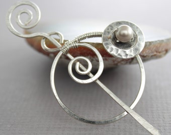 May include: A silver wire brooch featuring a circle and two spirals. A small white pearl is set in the centre of the circle.