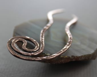 May include: A copper hair stick with a hammered finish and a spiral design at one end. The stick is resting on a piece of agate.