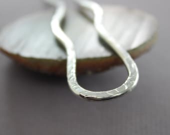 May include: Close-up of a silver hair pin with a hammered texture. The pin has a wavy design and is set against a blurred, neutral background. The metal has a bright, reflective quality.