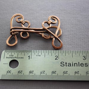 May include: A copper wire clasp with an intricate design, measuring approximately 2 inches in length. The clasp is shown next to a ruler for scale.