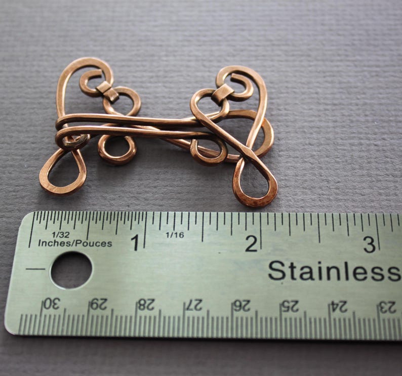 May include: A copper wire clasp with an intricate design, measuring approximately 2 inches in length. The clasp is shown next to a ruler for scale.