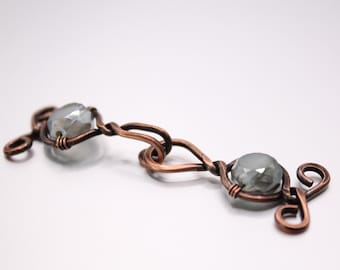 May include: A handcrafted copper wire bracelet with two light grey, faceted beads. The beads are held in place by the copper wire, which is shaped into decorative links. The bracelet has a rustic, artisan aesthetic, with a focus on the metalwork.