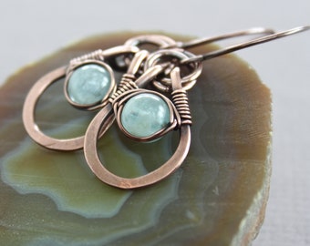 May include: A pair of copper wire wrapped earrings with light blue aquamarine stones. The earrings are hanging from a light brown agate slice.