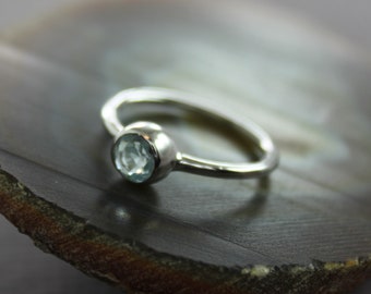 May include: A silver ring featuring a round, pale blue gemstone. The ring has a smooth, polished band, and the gemstone is set in a bezel. The ring is presented on a dark, textured surface.