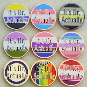 Può includere: Una collezione di 12 spille rotonde in smalto con la frase "It's Dr. Actually" in lettere dorate su una varietà di sfondi colorati. Ogni spilla presenta un diverso design di bandiera pride, tra cui la bandiera transgender, la bandiera lesbica, la bandiera bisessuale, la bandiera pansexuale, la bandiera genderqueer, la bandiera non binaria, la bandiera asessuale, la bandiera aromantica, la bandiera intersessuale, la bandiera poliamorosa e la bandiera genderfluid.
