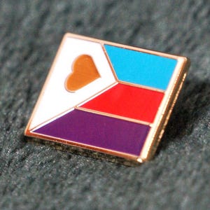 Op de afbeelding: Vierkante emaille pin met een gouden rand. De pin heeft een wit gedeelte met een bruin hart, een blauw gedeelte, een rood gedeelte en een paars gedeelte. De pin ligt op een donkergrijze, getextureerde ondergrond.