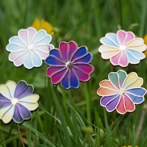 以下が含まれることがあります： 金色の縁取りが付いた、6つの花型のエナメルピンが緑の草の中に配置されています。ピンは、青、ピンク、紫、虹色のグラデーションカラーです。黄色の野花も確認できます。