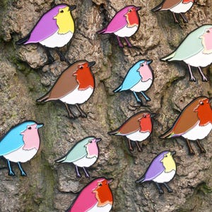 May include: A selection of bird-shaped enamel pins in various colours, including pink, blue, and brown, attached to a textured tree trunk. The pins feature white detailing and are designed to resemble robins.