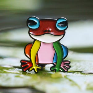 May include: A colorful frog pin with a red head, blue eyes, and a pink belly. The frog's body is a rainbow of colors, including yellow, orange, and green. The pin is set on a green lily pad, with its reflection visible in the water.