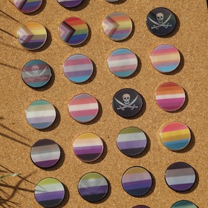 May include: A collection of round, colorful enamel pins featuring various LGBTQ+ pride flags and a skull and crossbones design. The pins are arranged on a corkboard, showcasing a variety of rainbow and striped patterns.