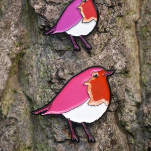 May include: Two robin enamel pins with a pink and red colour scheme. The birds have white bellies and are set against a textured tree bark background. The pins are a charming accessory.