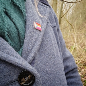 A woman with pink hair in a grey wool coat with a green scarf, wearing a rectangular rose gold pin with the trans chevron on the left and lesbian pride stripes.