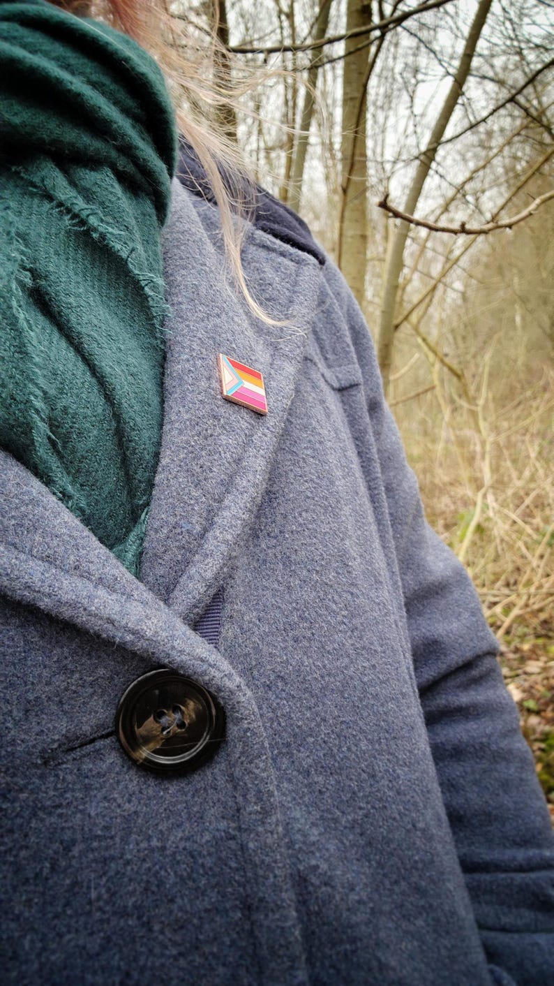 A woman with pink hair in a grey wool coat with a green scarf, wearing a rectangular rose gold pin with the trans chevron on the left and lesbian pride stripes.