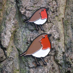 May include: Two enamel pins of a robin bird. Each pin has a brown back, white chest, and a red breast. The pins are set against a textured tree bark background. The birds are facing the same direction.