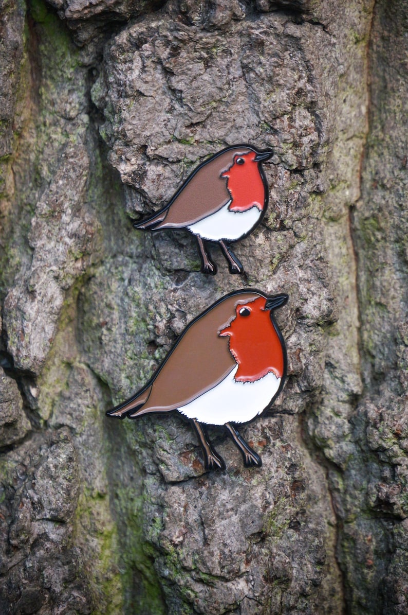 May include: Two enamel pins of a robin bird. Each pin has a brown back, white chest, and a red breast. The pins are set against a textured tree bark background. The birds are facing the same direction.