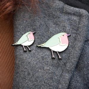May include: Two bird enamel pins. The pins feature a robin design with a light blue body, a pink breast, and black legs. The pins are displayed on a grey fabric background.
