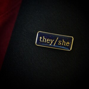 The they/she badge on a lapel. The background looks almost black with a hint of blue. The flecks of glitter in it look like stars
