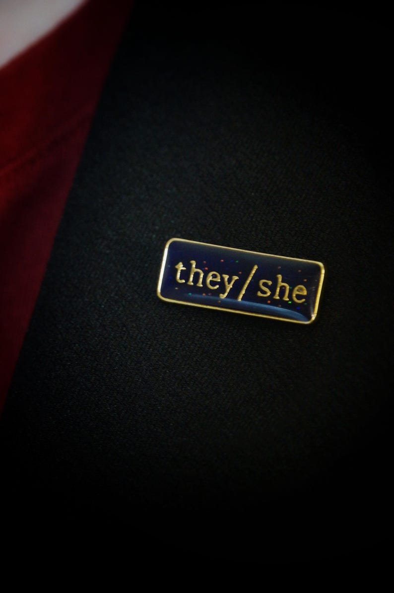 The they/she badge on a lapel. The background looks almost black with a hint of blue. The flecks of glitter in it look like stars