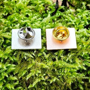 The backs of two rectangular pins with butterfly style clasps. One is silver and one is rose gold. The clasps are silver and gold respectively.