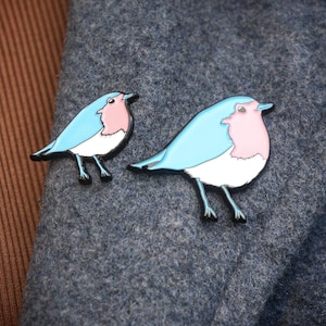 May include: Two bird-shaped enamel pins. Each pin has a light blue body, a pink breast, and a white belly, with black outlines and legs. The pins are displayed on a grey fabric.