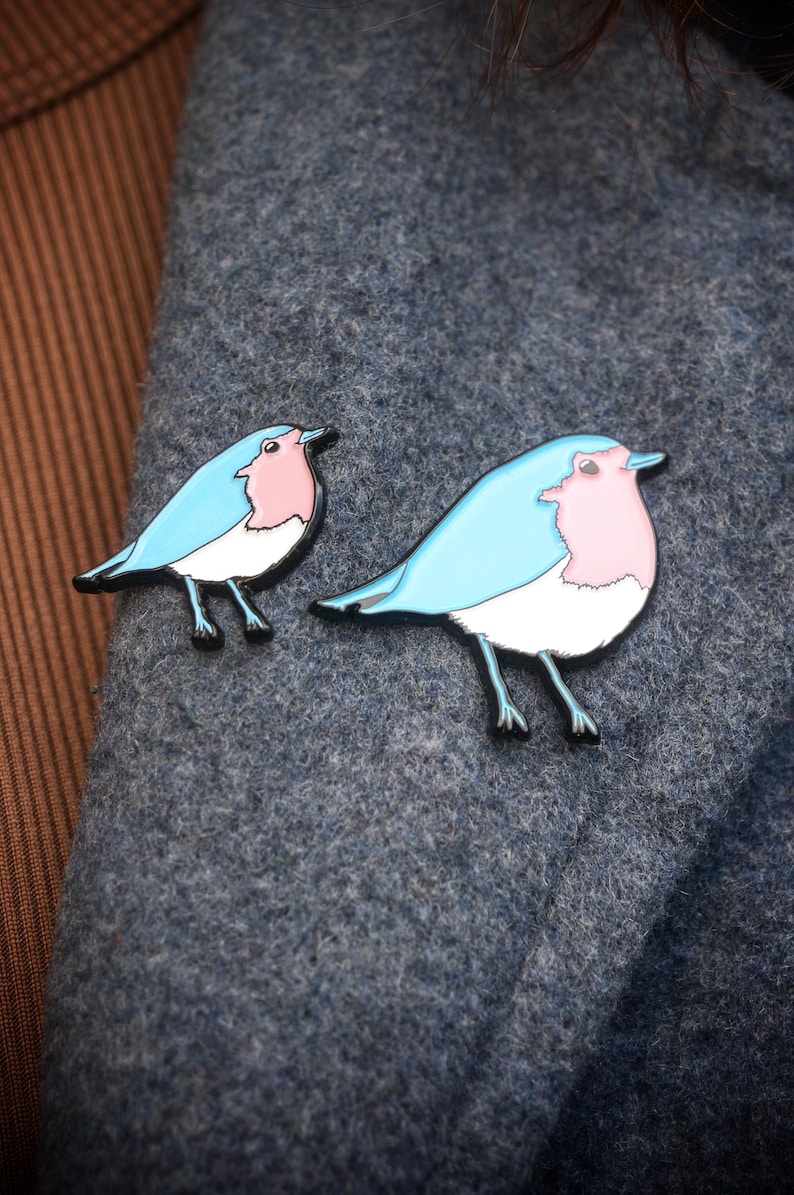 May include: Two bird-shaped enamel pins. Each pin has a light blue body, a pink breast, and a white belly, with black outlines and legs. The pins are displayed on a grey fabric.
