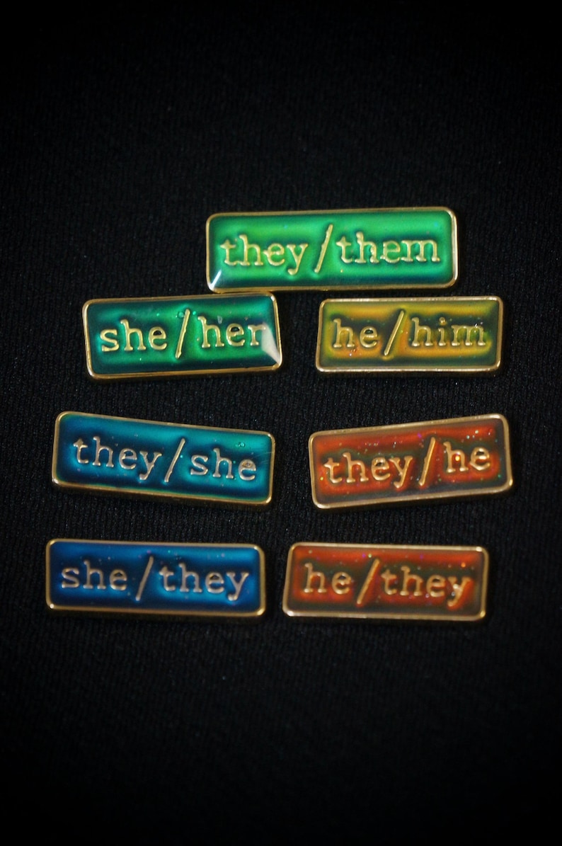 A collection of seven rectangular pronoun pins showing every combination of he, she, and they pronouns in gold text. The background colours are variously blue, greens, yellows, and oranges with small flecks of glitter.