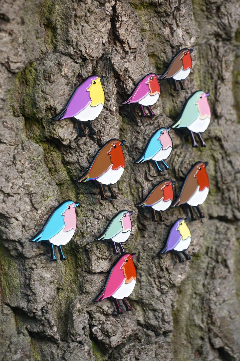 May include: A selection of bird-shaped enamel pins in various colours, including purple, pink, blue, brown, and green, attached to a tree trunk. The pins have white bellies and black legs. The birds have a cartoon design.