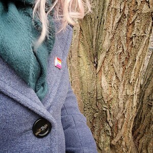 A woman with pink hair in a grey wool coat with a green scarf, wearing a rectangular rose gold pin with the trans chevron on the left and lesbian pride stripes.