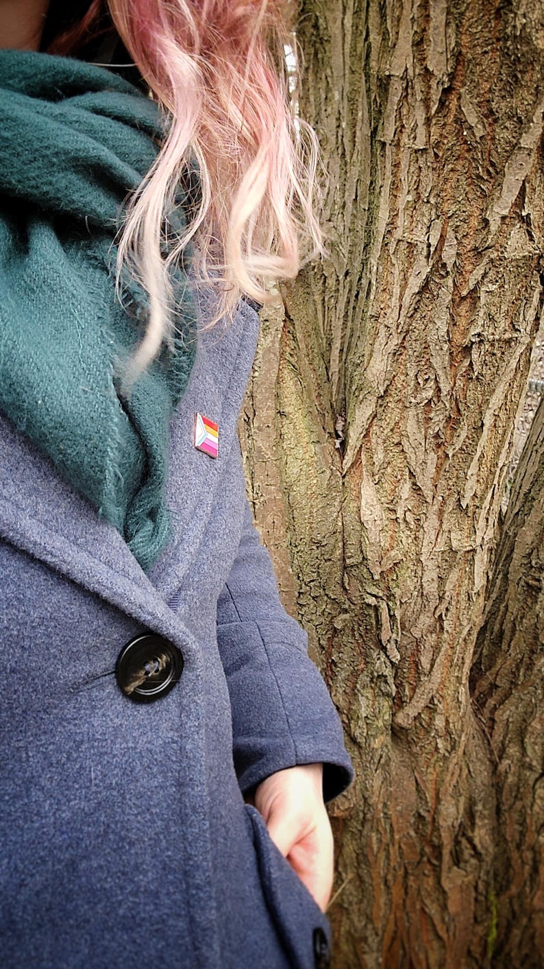 A woman with pink hair in a grey wool coat with a green scarf, wearing a rectangular rose gold pin with the trans chevron on the left and lesbian pride stripes.