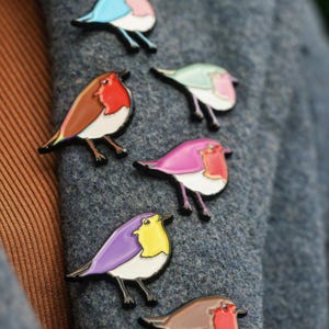May include: A selection of robin bird enamel pins in various colours, including blue, red, purple, and green, with white bellies and black outlines. The pins are attached to a grey wool coat.