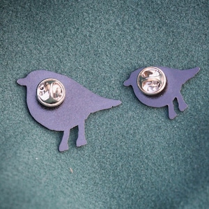 May include: Two dark blue bird-shaped enamel pins with gold-coloured pin backs. The pins are in different sizes, with one larger than the other. The pins are on a dark green textured surface.