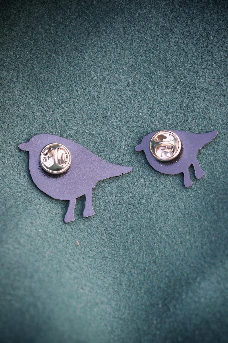 May include: Two dark blue bird-shaped enamel pins with gold-coloured pin backs. The pins are in different sizes, with one larger than the other. The pins are on a dark green textured surface.