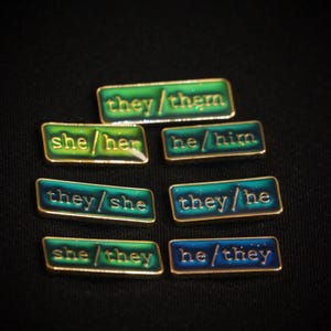 A collection of seven rectangular pronoun pins showing every combination of he, she, and they pronouns in gold text. The background colours are variously blue and greens with small flecks of glitter.